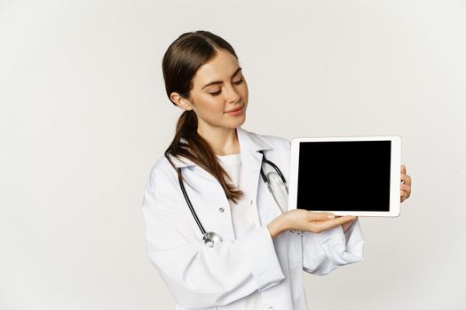 Image of woman doctor, female healthcare worker showing online medical website, digital tablet screen and smiling, standing in white coat over white background.