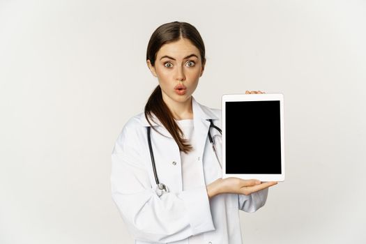 Image of woman doctor, female healthcare worker showing online medical website, digital tablet screen and smiling, standing in white coat over white background.