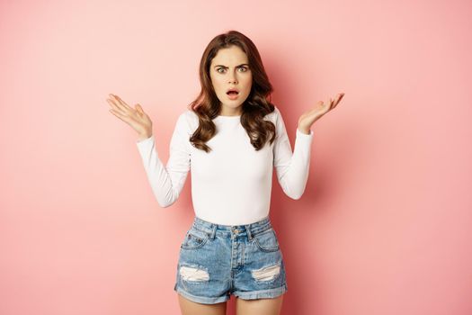 Frustrated young woman, staring at camera, oh my gosh face, frowning shocked, standing against pink background.