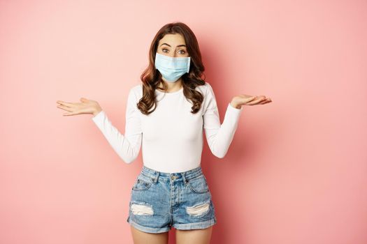 Covid-19. Confused young woman in medical face mask, shrugging shoulders and looking clueless, standing puzzled against pink background.