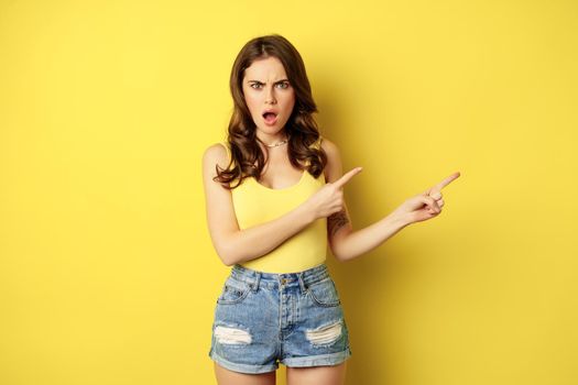 Shocked and frustrated young woman pointing right, showing banner or logo with angry, confused face expression, standing over yellow background.
