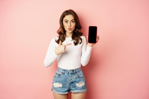 Surprised brunette girl pointing at mobile phone app screen, showing smartphone advertisement, online shopping offer, standing amazed against pink background.