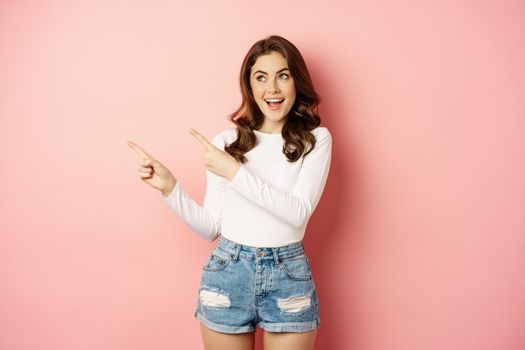 Cute smiling brunette girl, showing advertisement, pointing fingers left at banner, logo on empty pink space, studio background.