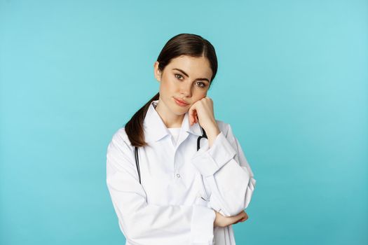 Thoughtful medical worker, young doctor woman looking unamused, bored, standing in white coat against blue background.