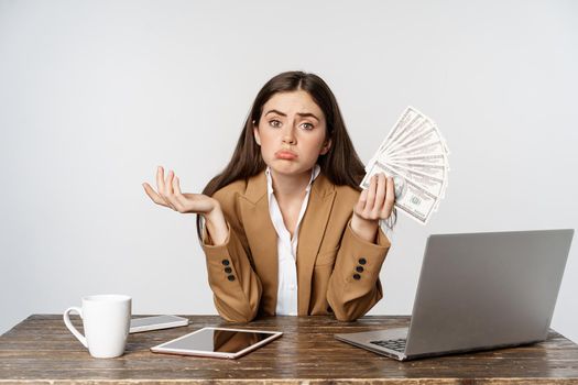 Sad businesswoman sitting in office with laptop, holding money and looking disappointed in income, business failure, white background.