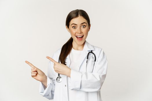 Portrait of smiling young woman doctor, healthcare medical worker, pointing fingers left, showing clinic promo, logo or banner, standing over white background.