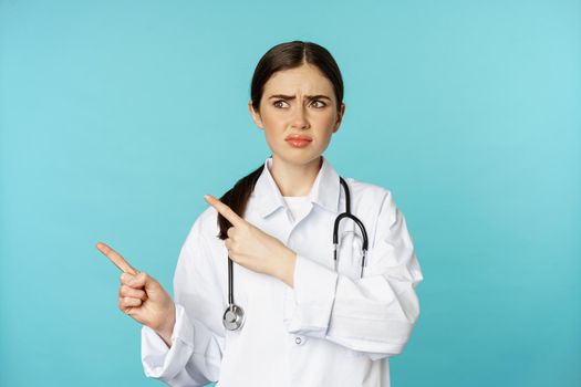 Confused, disappointed medical worker, frowning while pointing fingers left with doubtful, puzzled face expression, standing over torquoise background.