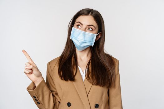 Coronavirus and people concept. Portrait of business woman at workplace wearing face mask, pointing finger left at logo, company banner, white background.