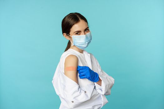 Young female doctor, nurse in medical face mask and hospital uniform, showing thumbs up after getting covid-19 omicron vaccine, showing patch on shoulder, blue background.