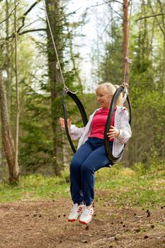 Middle aged woman in the woods swinging on a swing made from an old car tire. Blonde girl resting in nature