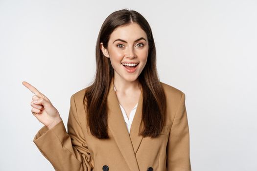 Portrait of businesswoman pointing finger left, showing corporate banner, logo, standing in brown suit over white background.