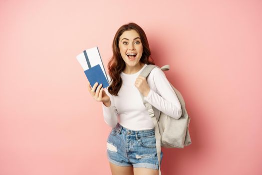 Vacation and holiday. Beautiful girl going on trip, holding passport with airplane tickets, holding backpack, travelling, standing over pink background.