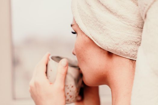 Young serene relaxed woman in spa bath towel drinking hot beverage tea coffee after taking shower bath at home. Beauty treatment, hydration concept.