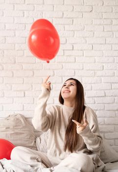 Valentine's day, Women's day. Young brunette woman sitting in the bed celebrating valentine day holding red heart balloons