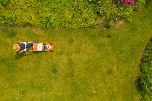 A young man is mowing a lawn with a lawn mower in his beautiful green floral summer garden. A professional gardener with a lawnmower cares for the grass, view from above