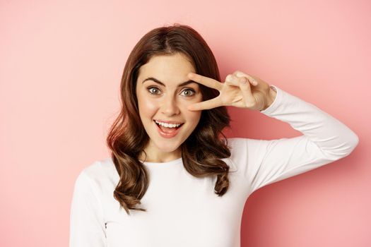 Close up portrait of coquettish young woman smiling, showing peace, v-sign gesture and posing happy, standing against pink background.