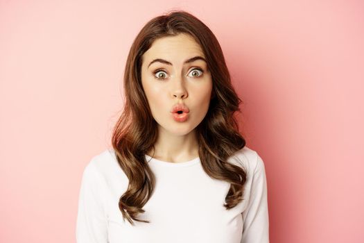 Image of stylish glamour woman looking surprised and amazed, saying wow with impressed reaction, standing over pink background.