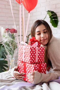 Valentine's day, Women's day. Young caucasian brunette woman sitting in the bed celebrating valentine day embracing pile of gifts