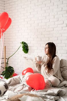 Valentine's day, Women's day. Young brunette woman sitting in the bed celebrating valentine day holding cup of coffee