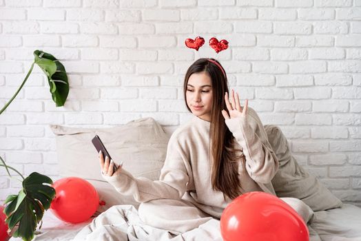 Valentine's day, Women's day. Young funny woman sitting in the bed celebrating valentine day chatting using mobile phone