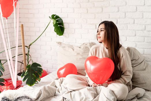 Valentine's day, Women's day. Young brunette woman sitting in the bed celebrating valentine day holding red heart balloons