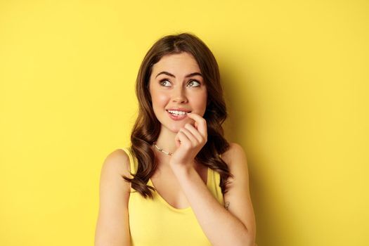 Close up portrait of stylish young beautiful woman thinking, yearning smth, looking up and making choice, standing over yellow background.