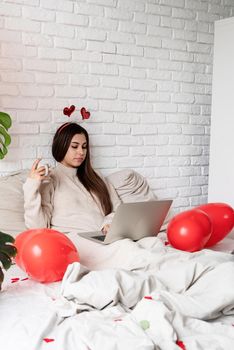 Valentine's day, Women's day. Young caucasian brunette woman sitting in the bed celebrating valentine day working on laptop online