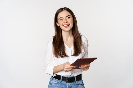 Smiling modern woman standing with digital tablet, laughing and looking happy, working, posing against white studio background.
