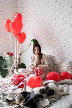 Valentine's day, Women's day. Young caucasian brunette woman sitting in the bed celebrating valentine day wrapping the gifts