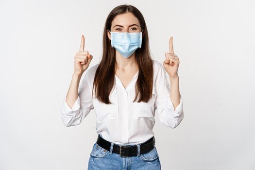 Portrait of corporate woman in face medical mask from coronavirus, pointing fingers up, showing advertisement, white background.