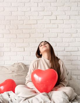 Valentine's day, Women's day. Young brunette woman sitting in the bed celebrating valentine day holding red heart balloons