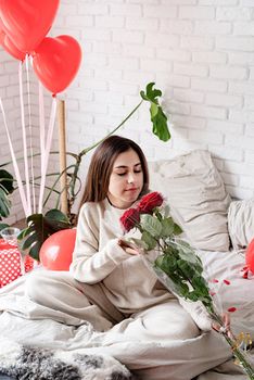 Valentine's day, Women's day. Young caucasian brunette woman sitting in the bed celebrating valentine day smelling red roses