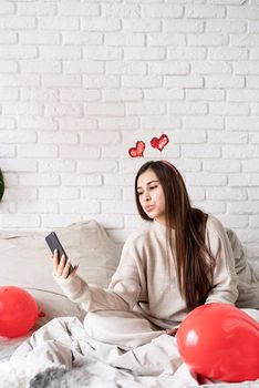 Valentine's day, Women's day. Young funny woman sitting in the bed celebrating valentine day chatting using mobile phone