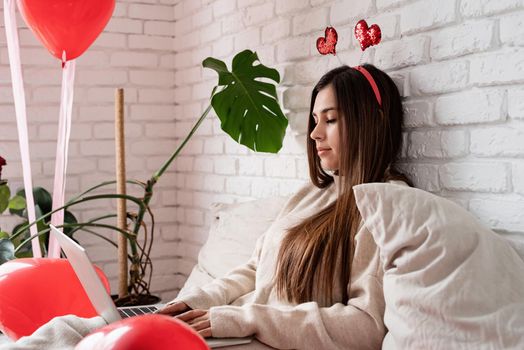 Valentine's day, Women's day. Young caucasian brunette woman sitting in the bed celebrating valentine day working on laptop online