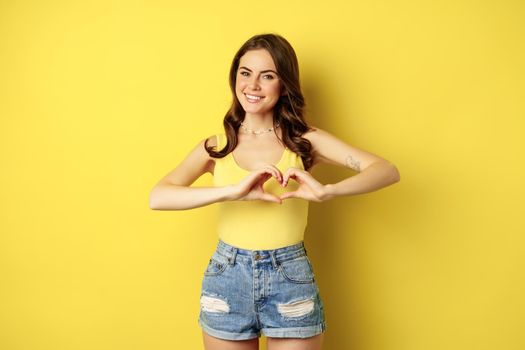 Beautiful young woman showing heart, love sign, like gesture, smiling happy and cute, standing in tank top and shorts over yellow background.