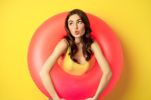 Close up portrait of coquettish young sexy woman inside pink swimming ring, concept of beach holiday, vacation and summer, yellow background.