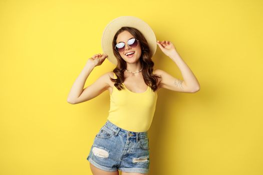 Stylish girl in straw hat and tank top, ready for summer, going on vacation and smiling pleased, standing over yellow background.