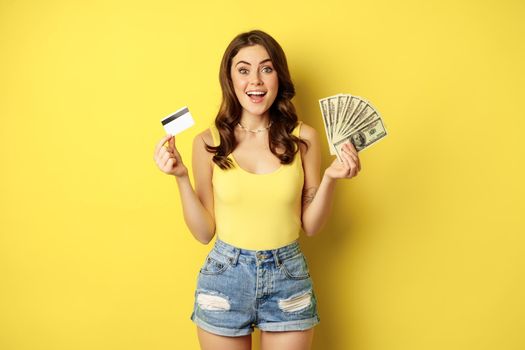 Young pretty woman in summer outfit holding credit card and money, cash in hands, standing against yellow background.