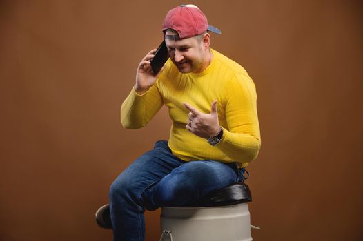 A man speaks expressively on the phone while sitting on a barrel in the studio. Chubby man talking on his smartphone emotionally in the studio.