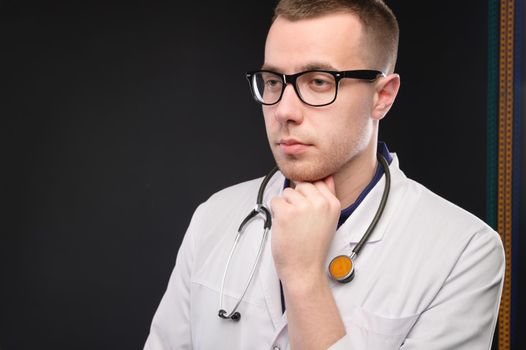 A young attractive Caucasian doctor in glasses and a white coat stands pensive and looks to the side. Studio portrait of a paramedic.