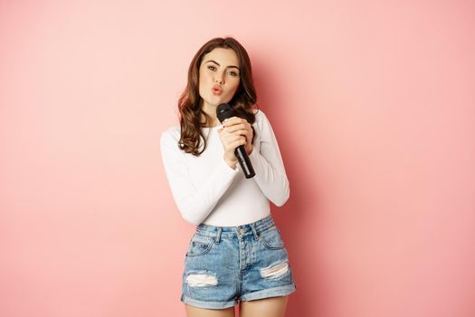 Karaoke. Happy smiling girl singing in microphone, dancing and having fun, standing over pink background. Events and party concept