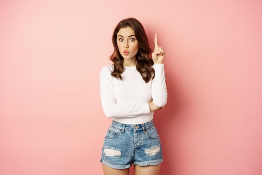 Young brunette glam girl raising finger, pointing up, showing promo offer, eureka gesture, realise something, standing over pink background.