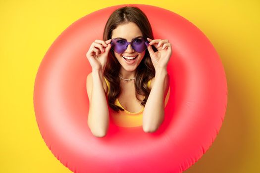 Close up portrait of stylish young woman, girl with swimming ring and summer sunglasses, ready for beach holiday, vacation, standing over yellow background.