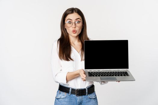 Portrait of cute girl in glasses, student showing laptop screen with amazed face, standing over white background. Copy space