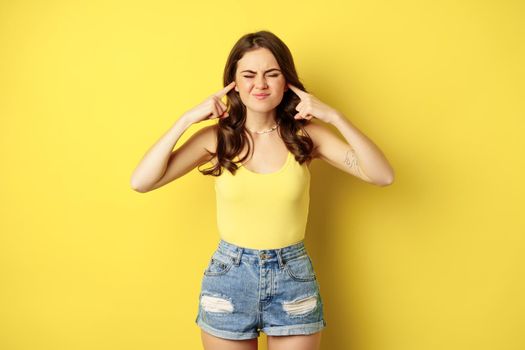 Young stylish woman feeling discomfort from loud noise sound, shut ears with fingers and complaining, standing over yellow background. Copy space