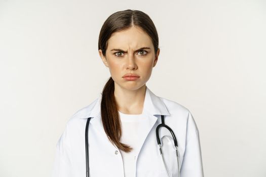 Angry and upset young woman doctor, female healthcare worker sulking, frowning disappointed, dislike smth, standing over white background.