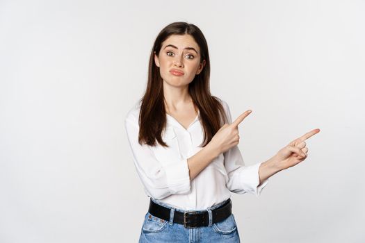 Sad and disappointed cute woman, gloomy girl pointing fingers at logo, complaining, sulking upset, standing in blouse over white background.