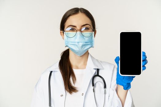 Online medical help concept. Woman doctor in glasses and face mask, showing mobile phone screen, app interface or website for patients, standing over white background.