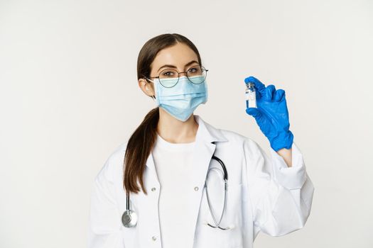 Portrait of young woman, doctor in medical face mask and uniform, showing vaccine, covid-19 vaccination campaign, standing over white background.