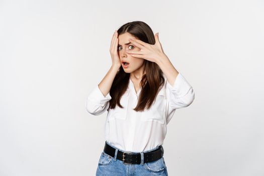 Intrigued, shocked young woman peeking through fingers at smth interesting, standing over white background.